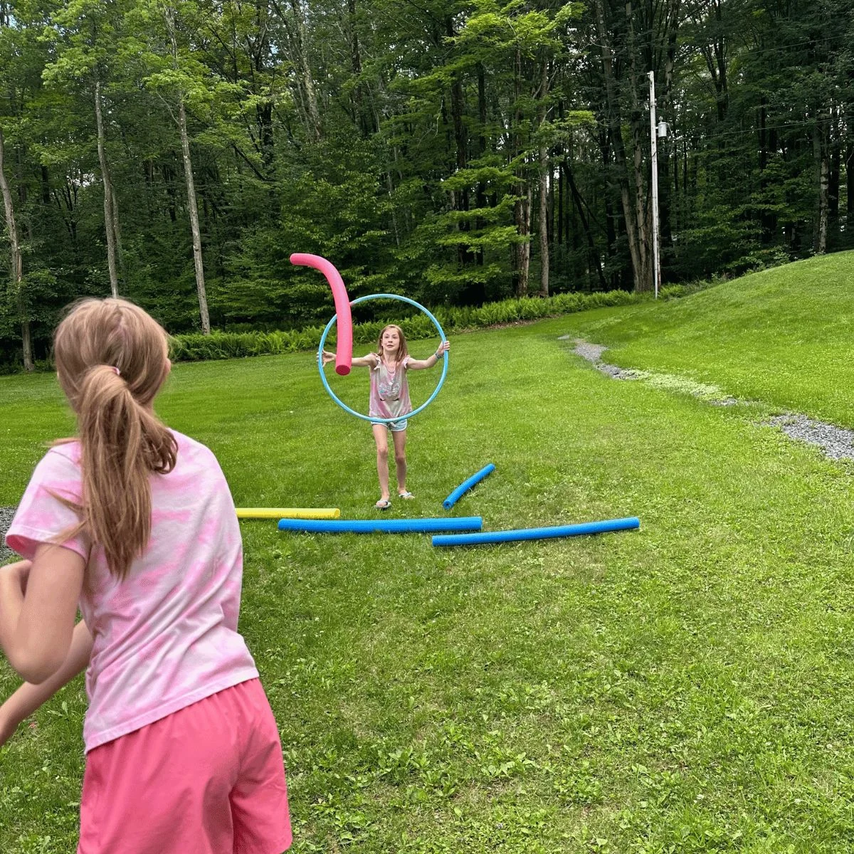 child trying to throw pool noodle through the hula hoop that her sister is holding